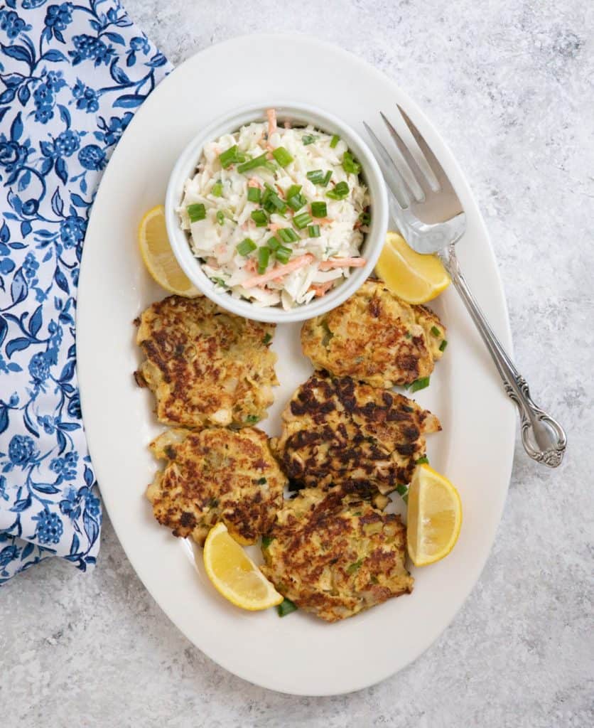 Chicken cakes on a white oval platter with a bowl of coleslaw, lemons and a spoon
