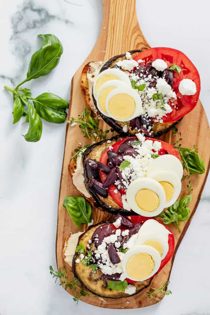 Eggplant and tomato slices with hummus on sliced bread topped with hard boiled eggs on a wooden board with basil garnish