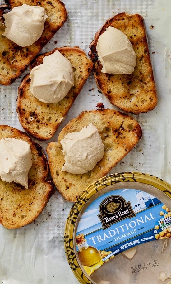 Slices of grilled bread on parchment paper topped with spoonfuls of Boarshead hummus
